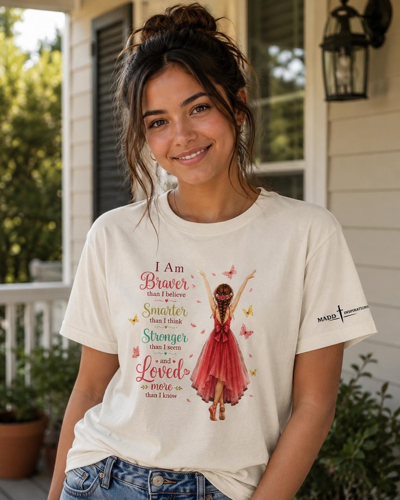 May include: A cream-colored short-sleeve t-shirt featuring a graphic of a person in a red dress with arms raised, surrounded by butterflies. The text reads: I Am Braver than I believe, Smarter than I think, Stronger than I seem and Loved more than I know.