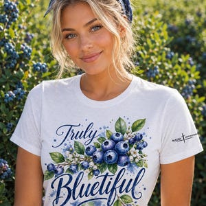 Op de afbeelding: Een wit T-shirt met korte mouwen en een grafische print van blauwe bosbessen, groene bladeren en witte bloemen. De tekst "Truly Bluetiful" is gedrukt in een donkerblauw scriptlettertype. De merknaam "MADD INSPIRATIONS" staat op de mouw.