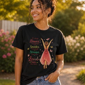 May include: A black short-sleeve t-shirt featuring a graphic of a person in a pink dress with arms raised, surrounded by butterflies. The text reads: I Am Braver than I believe, Smarter than I think, Stronger than I seem, and Loved more than I know.