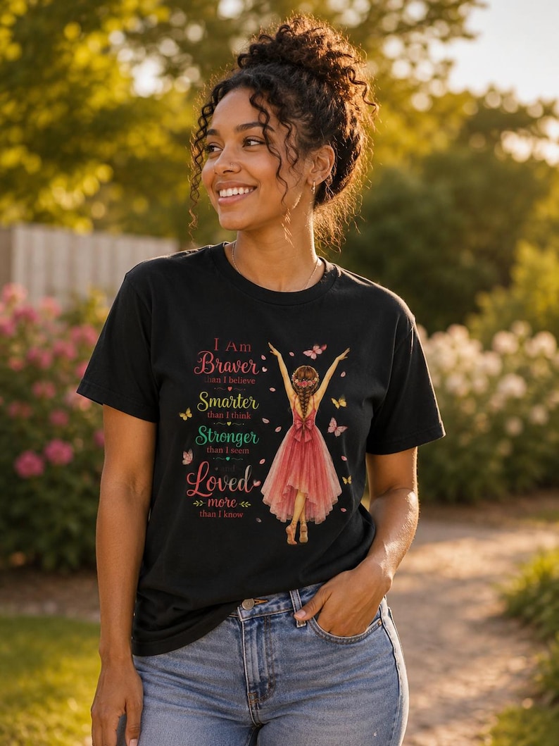 May include: A black short-sleeve t-shirt featuring a graphic of a person in a pink dress with arms raised, surrounded by butterflies. The text reads: I Am Braver than I believe, Smarter than I think, Stronger than I seem, and Loved more than I know.
