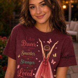 May include: A maroon short-sleeved t-shirt featuring a graphic of a person in a red dress with arms raised, surrounded by butterflies. The text reads: I Am Braver than I believe, Smarter than I think, Stronger than I seem and Loved more than I know.