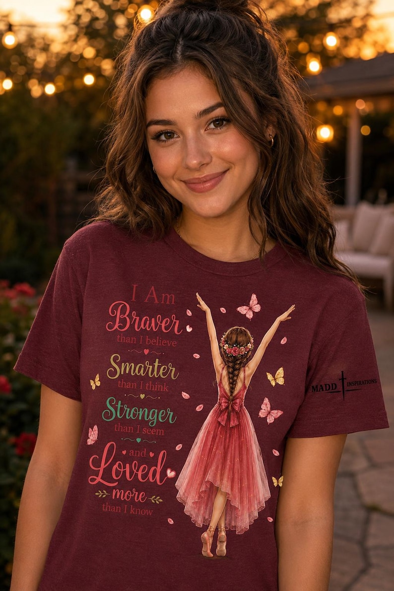 May include: A maroon short-sleeved t-shirt featuring a graphic of a person in a red dress with arms raised, surrounded by butterflies. The text reads: I Am Braver than I believe, Smarter than I think, Stronger than I seem and Loved more than I know.