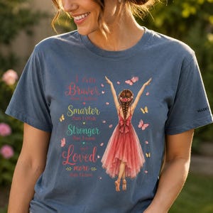May include: A blue short-sleeved t-shirt featuring a graphic of a person in a red dress with arms raised, surrounded by butterflies and petals. The text reads: I Am Braver than I believe, Smarter than I think, Stronger than I seem and Loved more than I know.