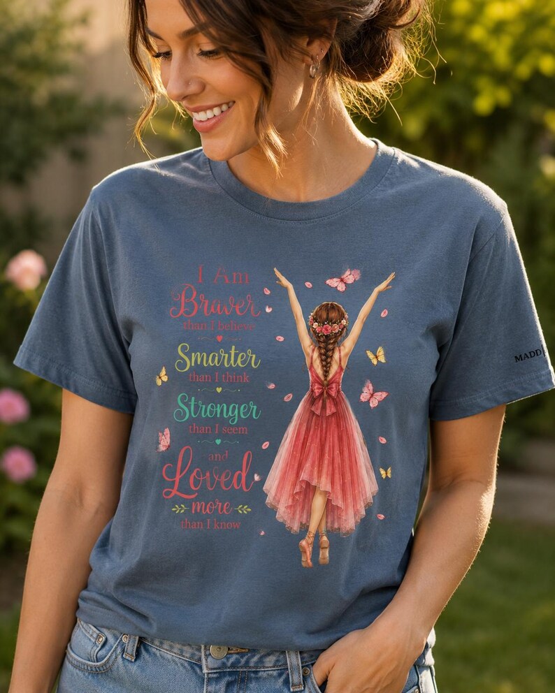 May include: A blue short-sleeved t-shirt featuring a graphic of a person in a red dress with arms raised, surrounded by butterflies and petals. The text reads: I Am Braver than I believe, Smarter than I think, Stronger than I seem and Loved more than I know.