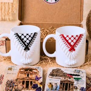 Pode incluir: Duas canecas de cerâmica branca com um padrão em relevo estilo keffiyeh em preto e vermelho com borlas brancas. Dois porta-copos quadrados exibem imagens impressas de Petra, Wadi Rum e do castelo de Ajloun numa caixa de presente de cartão.