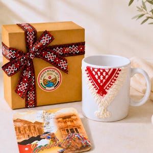 Pode incluir: Uma caixa de presente castanha com uma fita preta e vermelha, uma caneca branca com um design em relevo vermelho e branco estilo bordado, e um porta-copos quadrado com uma imagem de Petra e um camelo com o texto Jordan.