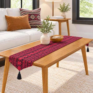 May include: A rectangular table runner featuring a vibrant red and black intricate floral pattern with black tassels attached to each end. The fabric displays a repeating symmetrical design that spans the length of the runner, providing a bold contrast.