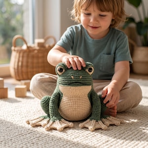 Könnte beinhalten: Ein handgefertigtes Häkel-Froschspielzeug mit dunkelgrünem Körper, cremefarbenem Bauch und Schwimmhäuten an den Füßen. Das Stofftier hat große, dunkle, runde Augen und einen gestickten Mund, was die detaillierte Garnstruktur des Designs hervorhebt.
