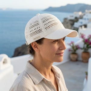 Può includere: Un cappellino da baseball bianco realizzato in filato all'uncinetto con una visiera strutturata e un motivo a trama aperta ventilata sulla corona. Una piccola etichetta rettangolare sul lato riporta il testo Handmade with Love in nero.