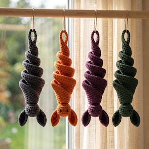 Op de afbeelding: Vier gehaakte vleermuisornamenten hangen aan een houten roede. Elke vleermuis heeft een spiraalvormig lichaam en twee oren, gemaakt van donkergrijs, oranje, paars en donkergroen garen. Elk figuurtje heeft twee kleine zwarte oogjes.