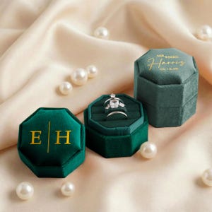 May include: Three dark green velvet ring boxes, one open with rings, and two closed. The boxes are octagonal with gold lettering. Pearls are scattered around the boxes on a cream-colored satin surface. The open box displays two rings.