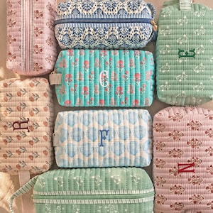 Op de afbeelding: Een collectie van zeven rechthoekige gewatteerde etuis met diverse bloemen- en schelpenpatronen in roze, blauwe en groene tinten. Verschillende etuis zijn geborduurd met individuele letters zoals C, E, F, N en R in contrasterend garen.