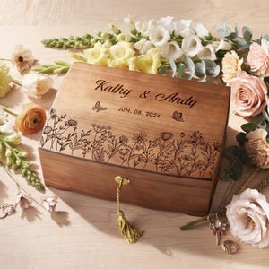 May include: A rectangular wooden keepsake box with a hinged lid featuring a laser-engraved floral meadow design, two butterflies, and the text Kathy & Andy JUN. 08, 2026. The box includes a gold-colored metal key with a gold tassel attached to the front.