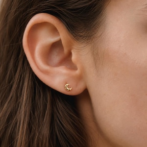May include: A gold-coloured crescent moon stud earring. The earring is small and detailed, with a curved shape and a textured surface. The earring is worn on an ear.