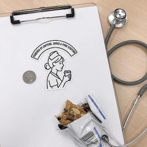 May include: A white clipboard with a sticker that reads "Powered by Caffeine, Chaos & Poor Staffing" featuring a nurse with a coffee cup. A stethoscope, a quarter, and a partially eaten granola bar are also present.