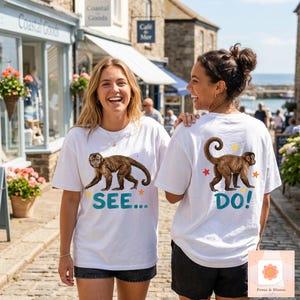 May include: Two people wearing white t-shirts with a brown monkey illustration. The front of one shirt says "SEE..." in teal, and the other says "DO!" The shirts have colorful star accents. The image is taken outdoors.