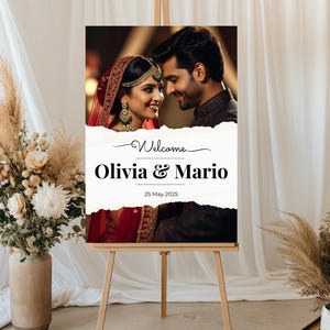 May include: A wedding welcome sign on a wooden easel. The sign features a photo of a smiling couple and the words "Welcome Olivia & Mario 25 May 2025" in black text. The background includes floral arrangements and white drapery.