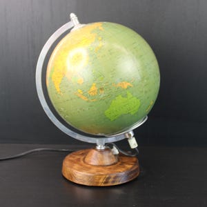 Puede incluir: Una lámpara de globo vintage con un diseño de mapa verde y amarillo. El globo está montado sobre un marco de metal plateado y una base de madera. La lámpara tiene un cable de alimentación.