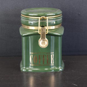 Può includere: Un contenitore in ceramica verde scuro con base quadrata e angoli arrotondati. Presenta un meccanismo di chiusura in metallo dorato sulla parte anteriore e la parola COFFEE stampata in lettere dorate sul pannello frontale. Il coperchio è fissato da una fascia metallica.
