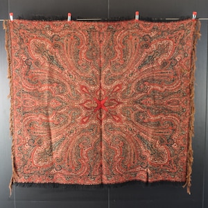 Peut inclure: Écharpe carrée à motif cachemire dans les tons de rouge, noir et beige. L'écharpe est bordée de franges et est suspendue à une corde à linge avec des pinces à linge rouges. Le tissu semble doux.