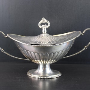 Op de afbeelding: Een zilverkleurige metalen schaal met deksel, voorzien van een geribbeld ontwerp, twee gebogen handgrepen aan de zijkanten en een voetstuk. Het deksel heeft een decoratieve knop en een geschulpte rand, terwijl de voet is afgewerkt met een touwachtig motief.