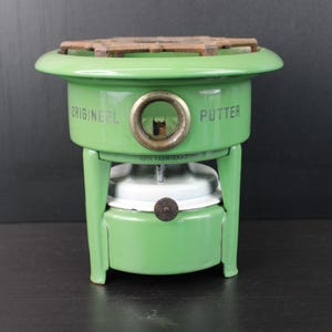 Op de afbeelding: Een vintage groene metalen kachel met de woorden "ORIGINEEL" en "PUTTER" aan de zijkant. De kachel heeft een ronde bovenkant met een bruin rooster en een messing ring. De kachel staat op een zwart oppervlak.