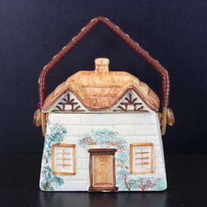 Puede incluir: Un recipiente de cerámica con forma de casa y tapa extraíble con diseño de techo de paja. La estructura tiene paredes de ladrillo blanco, una puerta central marrón, dos ventanas y un asa texturizada marrón unida a los lados del techo.