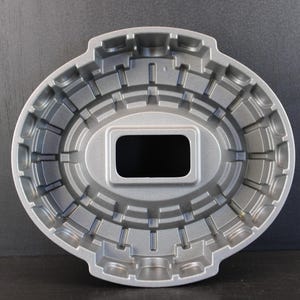 Könnte beinhalten: Eine silberfarbene Metall-Backform in ovaler Form mit einem stadionartigen Design. Das Innere weist eine Reihe von erhabenen rechteckigen Rippen und eine zentrale rechteckige Öffnung auf, die ein strukturiertes Muster für Kuchen erzeugt.