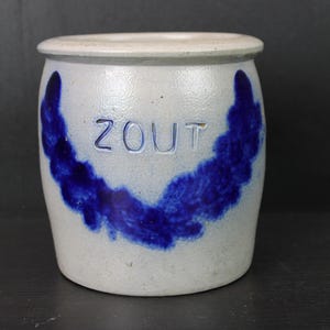 May include: A stoneware crock with a light gray body and a blue decorative design. The word "ZOUT" is inscribed in the center. The crock has a rounded shape and a slightly flared rim.