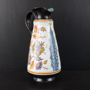 Könnte beinhalten: Ein Keramikkrug mit schwarzem Henkel, Rand und Boden. Der weiße Körper zeigt ein buntes Blumenmuster mit orangefarbenen, blauen, grünen und gelben Akzenten sowie ein vertikales blaues Paneel mit gelben Punkten und grünen Blattmotiven.