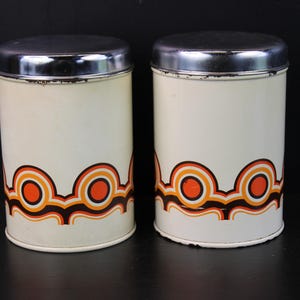 Op de afbeelding: Twee vintage witte metalen blikken met chromen deksels. Elk blik heeft een herhalend retro-ontwerp in oranje, bruin en crème. De blikken zijn cilindrisch en staan op een donkere ondergrond.