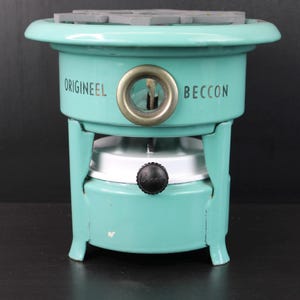 Può includere: Un fornello BECCON vintage color turchese con la scritta "ORIGINEEL" stampata sul lato. Il fornello ha una parte superiore rotonda con una superficie grigia, un anello di ottone e un pomello nero. Un classico degli utensili da cucina.