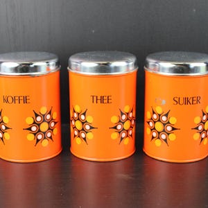 May include: Three orange metal canisters with silver lids. Each canister is labelled with a different word: "KOFFIE", "THEE", and "SUIKER". The canisters have a retro design with a repeating pattern of yellow, orange, and black shapes.