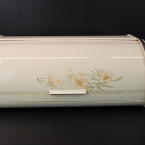 Peut inclure: Boîte à pain en métal crème avec un couvercle roulant et une garniture argentée. Le devant présente un motif floral jaune et vert. Une poignée blanche est centrée sur le devant. Conçu pour le stockage de la cuisine.