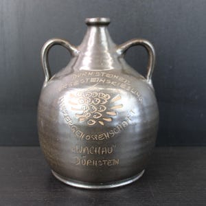 Può includere: Una brocca in ceramica marrone scuro con due manici e un collo stretto. La brocca presenta testo in rilievo e un emblema decorativo. Il testo include le parole "Dürnsteiner Winzergenossenschaft" e "Wachau Dürnstein".