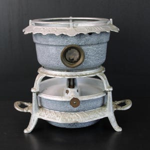 Könnte beinhalten: Ein Vintage-Fondue-Topf mit zweistufigem Design und einer gesprenkelten blau-grauen Oberfläche. Der Topf hat einen dekorativen, verzierten weißen Metallständer und Griffe. Der obere Rand hat eine dekorative Kante. Zum Erhitzen und Servieren von Fondue.