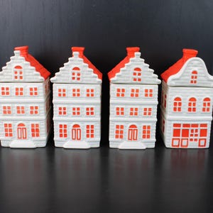 May include: A set of four ceramic canisters shaped like tall, narrow houses. Each canister is white with bright orange accents on the roof, windows, and doors. The structures feature stepped gables and detailed brick textures on the exterior walls.