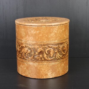 May include: A cylindrical, tan-coloured box with a lid. The box features a detailed, embossed band with animal and floral designs. The lid also has a subtle embossed pattern. The box is made of a textured material.