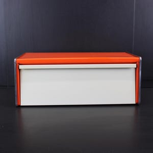 May include: A rectangular bread bin featuring a vibrant orange top lid and a solid off-white front panel. The storage container has a clean, boxy silhouette with metallic silver trim along the edges, providing a structured and functional design.