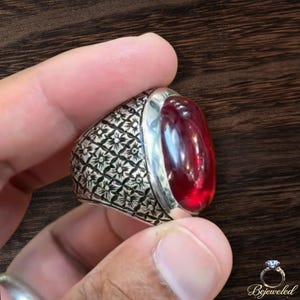 Op de afbeelding: Een zilveren ring met een grote, gladde, ovaalvormige dieprode edelsteen in een gepolijste metalen zetting. De ringband is breed en versierd met een herhalend kruislings patroon van kleine, gegraveerde stervormige motieven over het oppervlak.