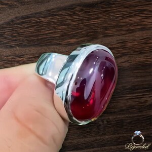 Op de afbeelding: Een zilveren ring met een grote, gladde, ovaalvormige dieprode edelsteen in een gepolijste metalen band. De steen heeft een hoogglanzende afwerking die licht reflecteert en het woord Bejeweled staat in de rechterbenedenhoek van de afbeelding.