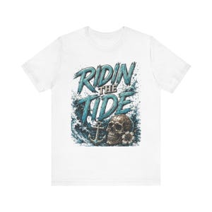 May include: White t-shirt with a graphic design featuring the text "RIDIN THE TIDE" in blue, over a skull, anchor, and ocean waves. The skull is detailed with a flower.