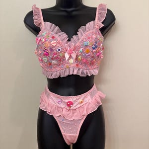 Puede incluir: Conjunto de lencería rosa sobre un maniquí negro. El sujetador y el tanga están adornados con cuentas, abalorios con forma de caramelo y volantes. El sujetador tiene tirantes con volantes y un lazo. El tanga tiene una cintura con volantes y un adorno de piruleta.