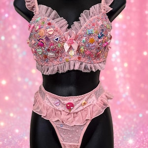 Peut inclure: Un ensemble de lingerie rose composé d'un soutien-gorge à volants et d'une culotte assortie. Le soutien-gorge est orné de breloques en forme de bonbons, de perles et d'un nœud central. Le tissu est agrémenté de strass et de volants en maille.