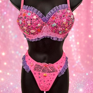 Peut inclure: Un ensemble soutien-gorge et culotte rose avec des bordures à volants violettes et des détails en dentelle. Le tissu est orné de nombreuses perles colorées, de petits charms en plastique en forme de bonbons et de cœurs avec l'inscription XOXO.