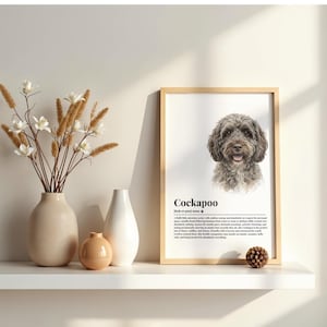 Puede incluir: Un cuadro con un retrato de perro Cockapoo, con el nombre de la raza y una descripción. La impresión se muestra en una estantería blanca con jarrones y flores secas. El marco es de madera clara.