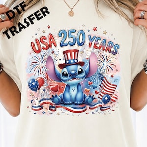 May include: A cream-coloured t-shirt featuring a graphic of a blue alien character wearing a red, white, and blue top hat. The design includes the text "USA 250 YEARS" surrounded by fireworks, American flag motifs, balloons, and hearts in red, white, and blue.