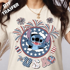 May include: A cream-coloured t-shirt featuring a graphic of the character Stitch inside a blue circle with white stars. The design includes red, white, and blue fireworks, two popsicles, a pinwheel, and the letters USA in patriotic patterns.