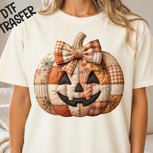 Peut inclure: Un t-shirt couleur crème arborant un grand motif central représentant une citrouille matelassée avec un visage de lanterne d'Halloween. La citrouille est faite de divers empiècements orange, marron et beige avec des toiles d'araignée.