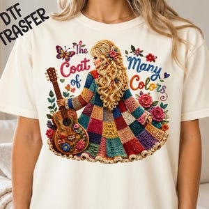 May include: A cream-colored t-shirt featuring a graphic of a person with long blonde hair wearing a multi-colored patchwork coat, holding an acoustic guitar. The text reads "The Coat of Many Colors" surrounded by colorful flowers and a butterfly.